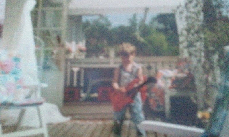 suddigt foto på mig när jag är kanske 3–5 år, på en veranda, med en leksaksgitarr i handen