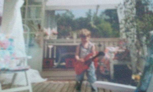 suddigt foto på mig när jag är kanske 3–5 år, på en veranda, med en leksaksgitarr i handen